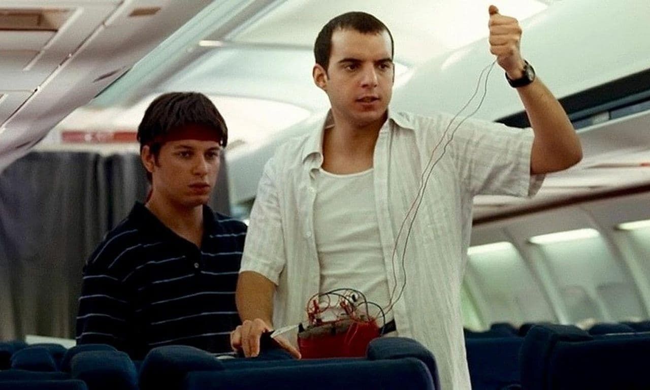 Two men standing in an airplane aisle, one holding up a thumb and the other watching nearby.