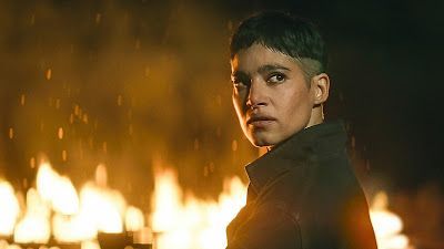 Woman with dark hair and a serious expression in front of a fire.