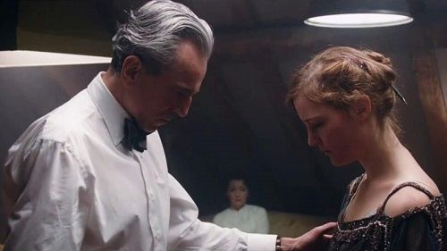 A man in a white shirt examines a woman's dress, both in a dimly lit room. Another woman watches.