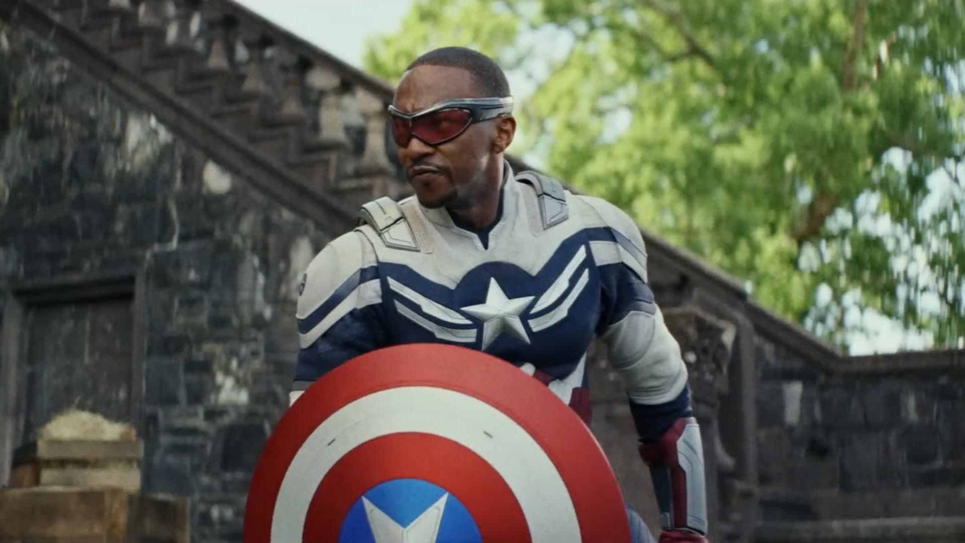 Captain America stands with his shield, wearing a blue suit and goggles in a stone outdoor setting.