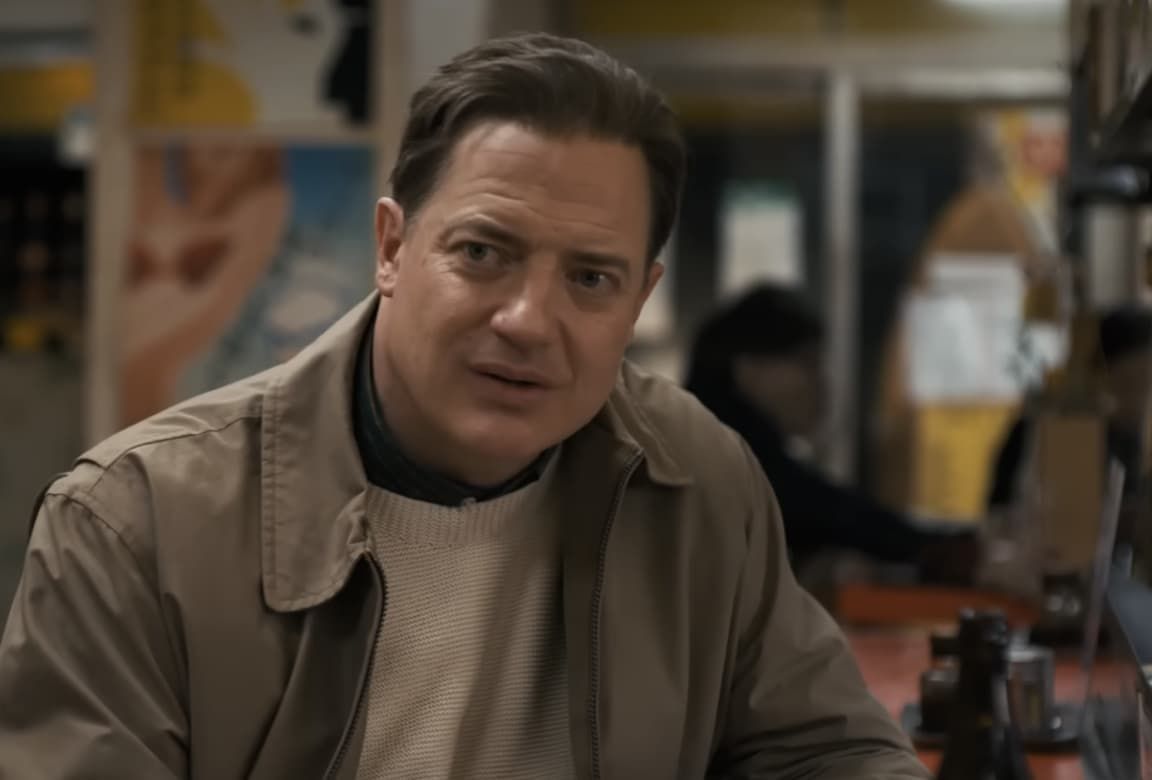 Man seated in a café, wearing a tan jacket and cream sweater, looking toward the camera.