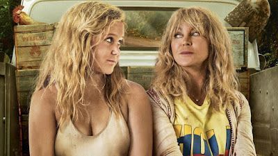 Two blonde women in a truck bed, looking at each other. One wears a beige top, the other a yellow shirt.