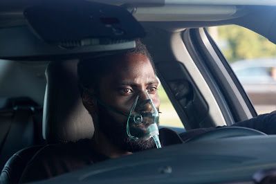 Driver wearing a clear oxygen mask in a car, looking ahead with a serious expression.