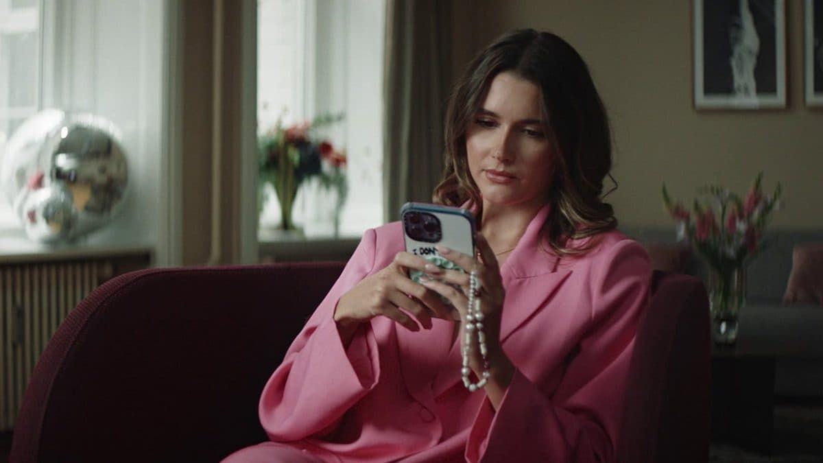 Woman in a pink suit smiling while holding a smartphone on a sofa in a dim living room.