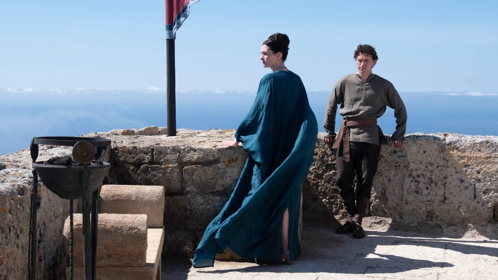 Two people in medieval-style clothing standing on a stone terrace with mountains in the background
