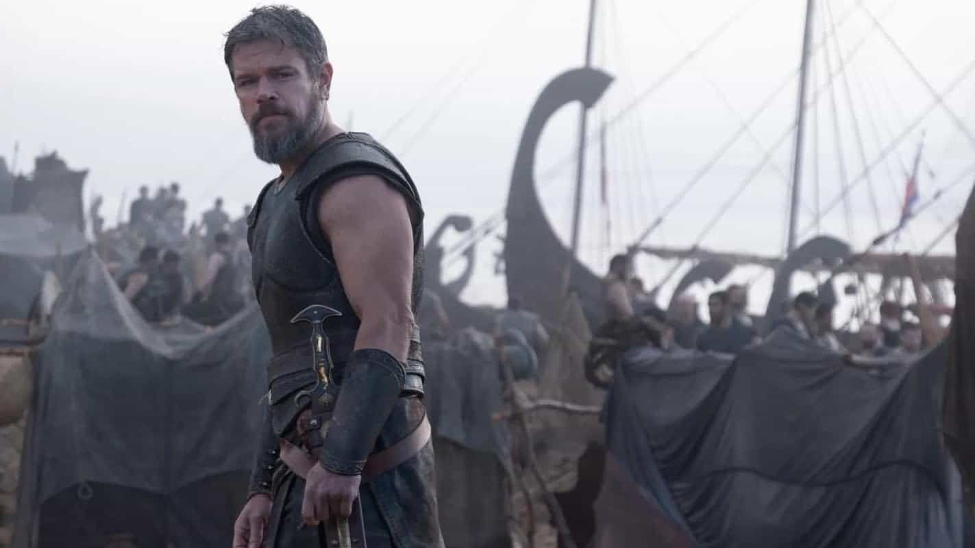A bearded warrior in leather armor stands on a ship deck with sails and shields in the background.