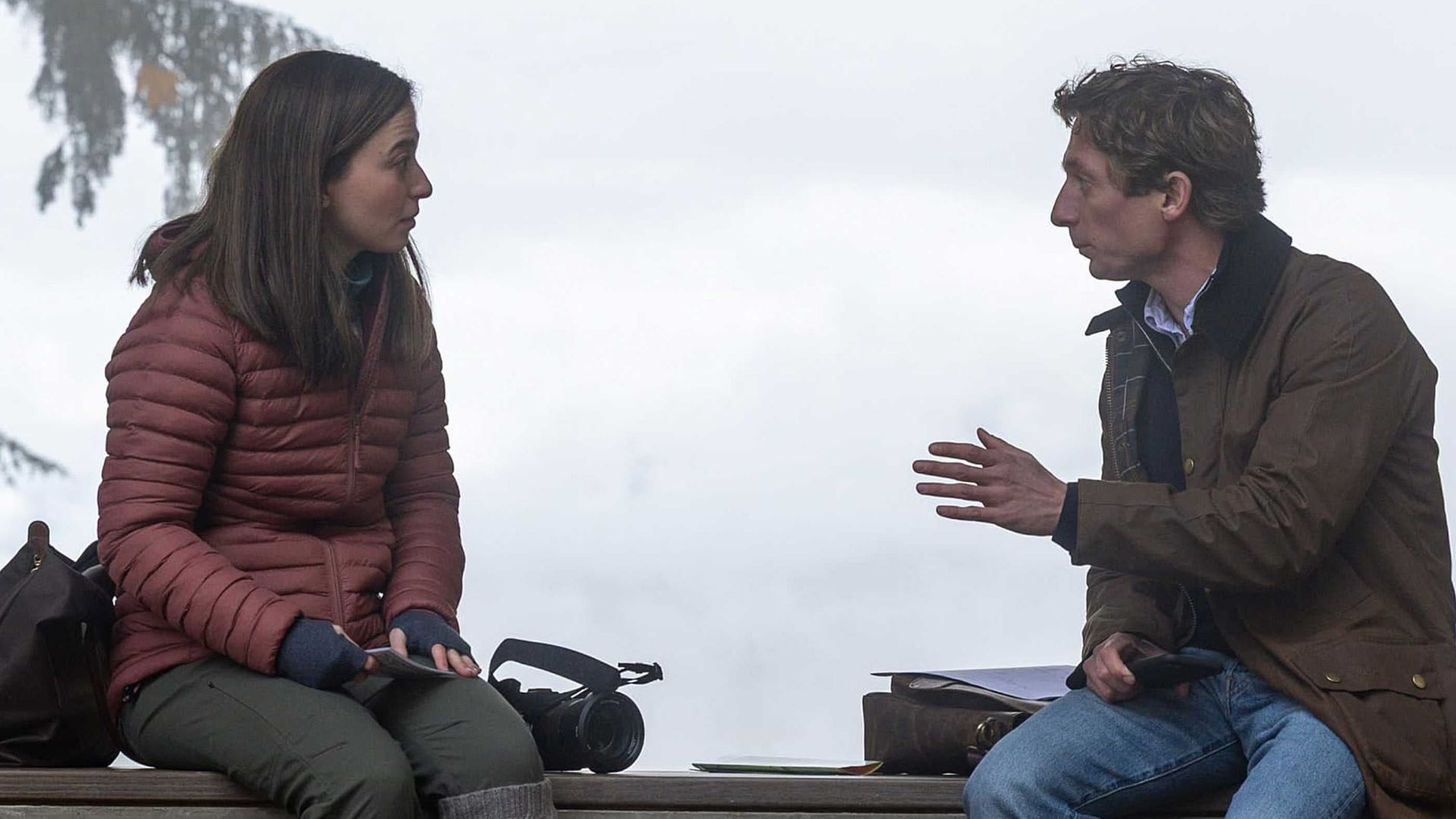 Two people sit outdoors on a bench, talking in a misty setting.