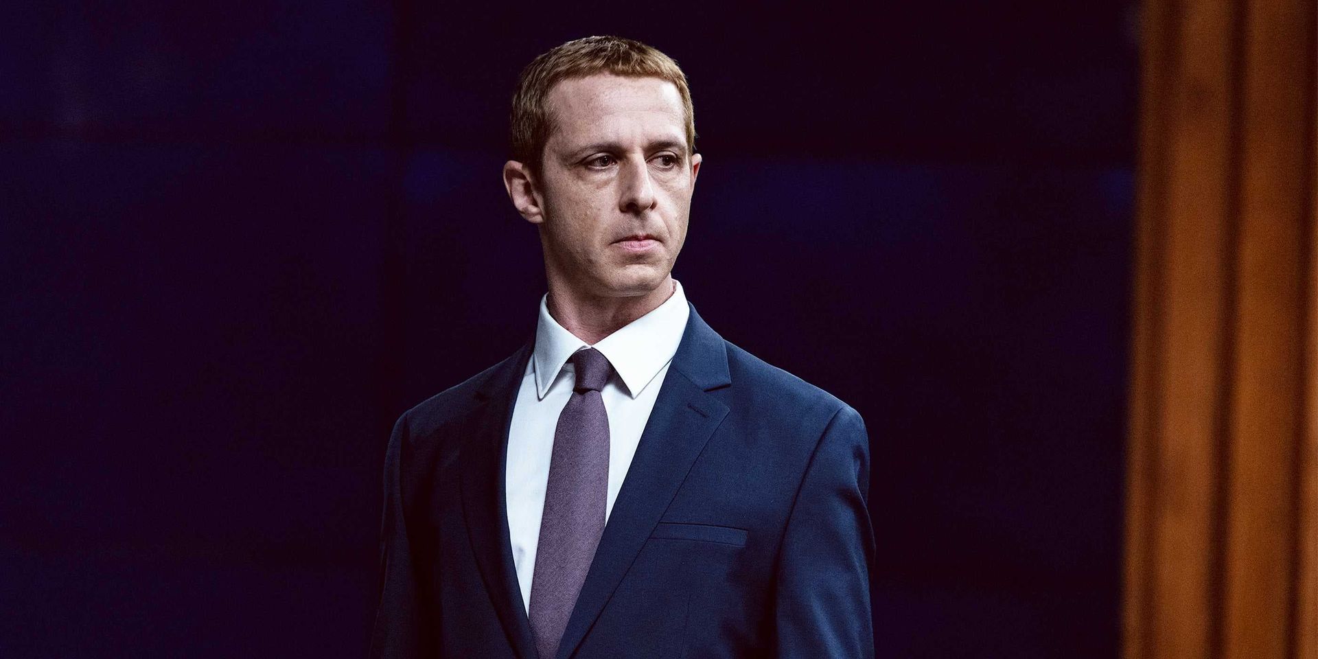 Man in a dark suit and tie standing against a dark stage background, looking to the side.