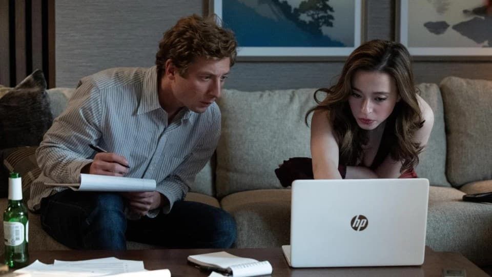 Two people on a couch reviewing notes beside an open laptop in a living room.
