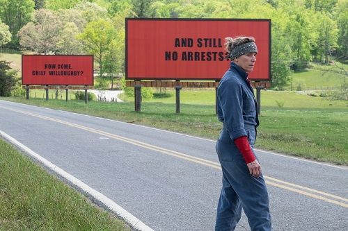Woman in jumpsuit by billboards on a road; billboards read