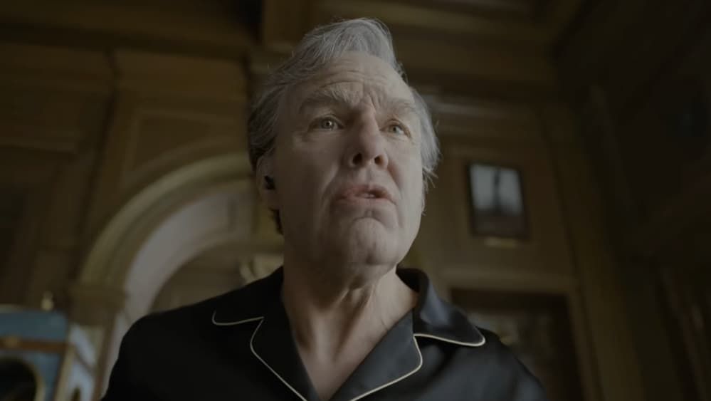 A stern person in a dark pajama top stands in a dim, ornate room, looking upward.