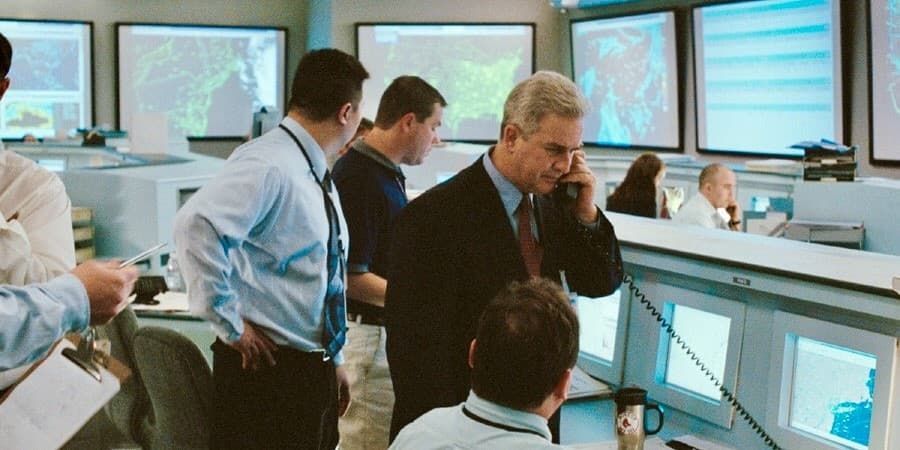 People gathered around monitors in a control room, discussing data on multiple screens.
