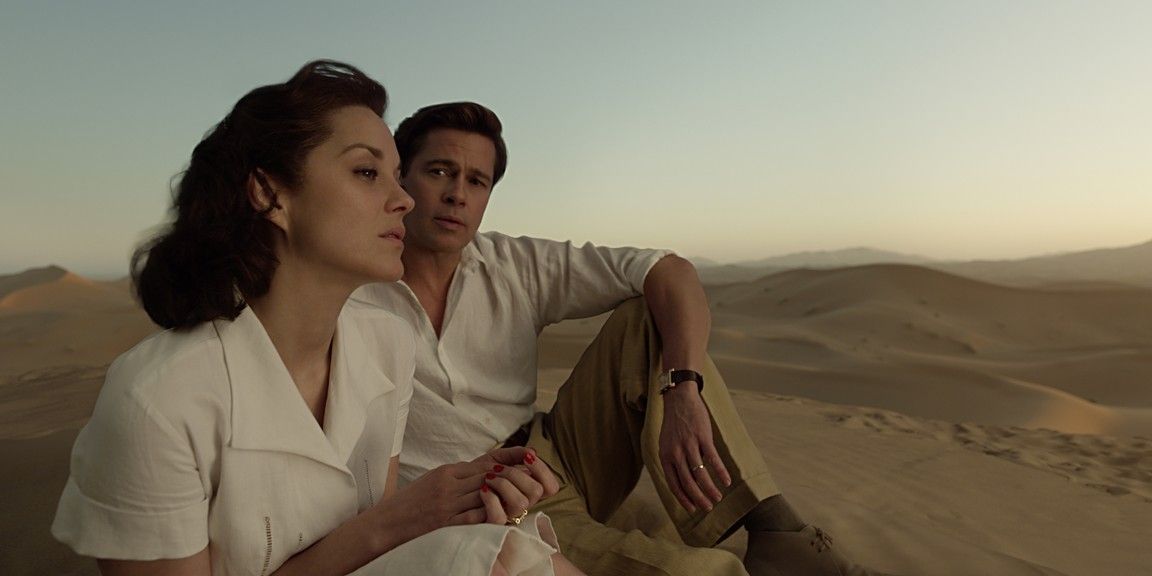 Man and woman sit on a sand dune, looking into the distance; both dressed in white/tan clothing.
