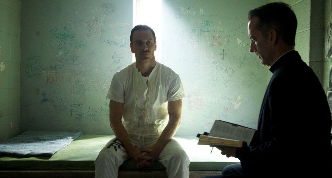 A man in a prison cell smiles at a priest reading from a book. Sunlight streams in from above.