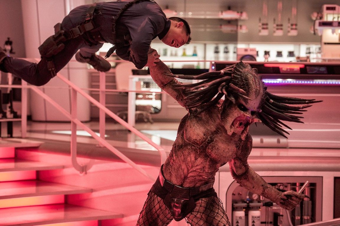 Predator fighting a person, both mid-air. Interior, red-lit room with stairs and a bar.