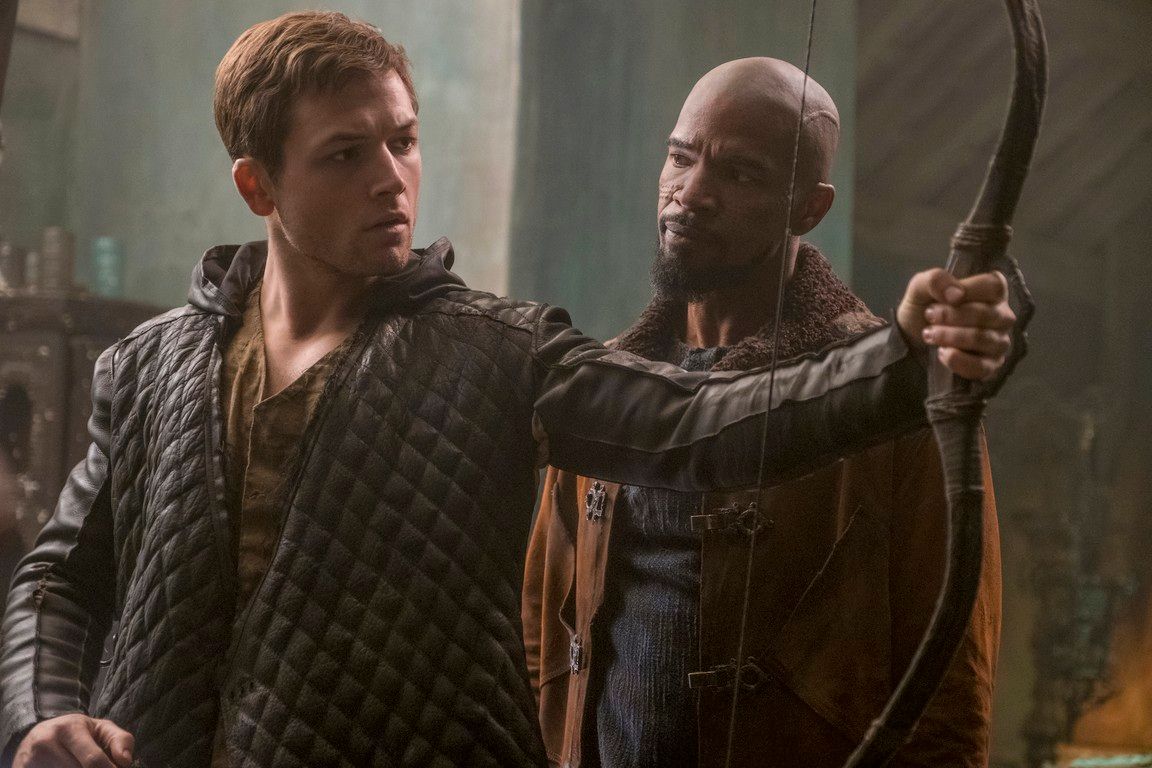 Robin Hood and John training with a bow and arrow in a dimly lit workshop.