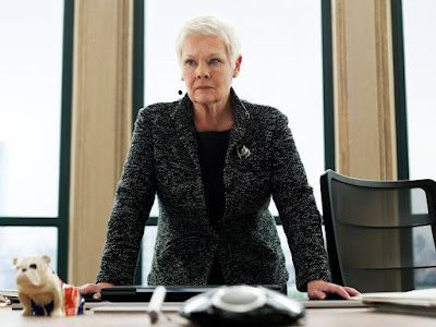 Judi Dench as M, sternly leaning on a desk in an office, a bulldog figurine sits nearby.