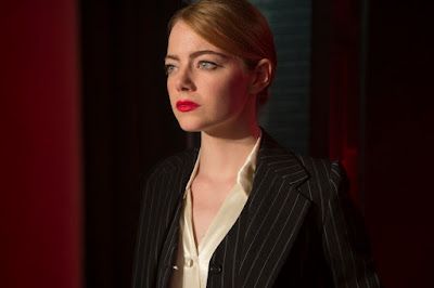 Woman in a black pinstripe blazer and white blouse, lit against a dark red background.