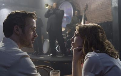 Two people talk at a dim bar while a singer performs on a small stage behind them.
