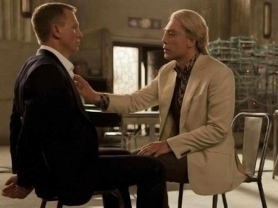 Bond and Silva in a dimly lit room, Silva touching Bond's chest; both dressed in suits, Bond bound.