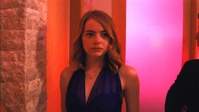 Woman in a blue dress standing in a pink-lit hallway near a stone wall.