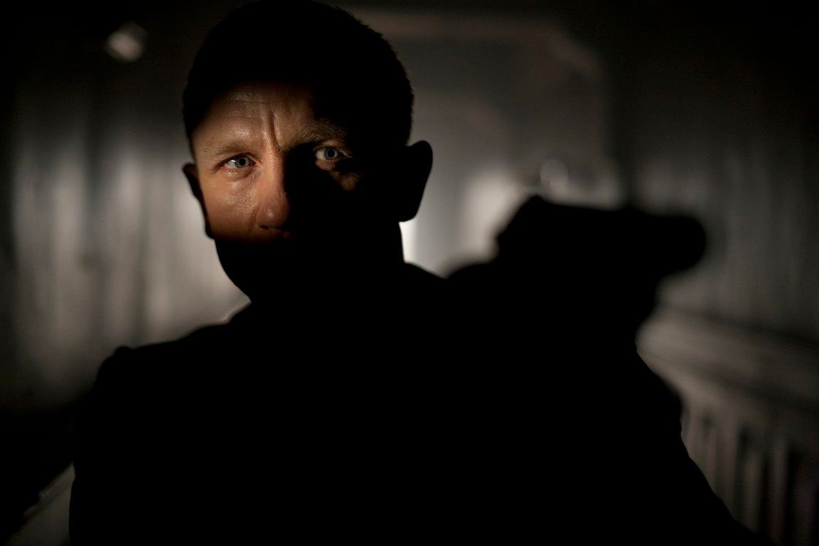Man in shadow aiming a gun down a dim hallway; serious expression.
