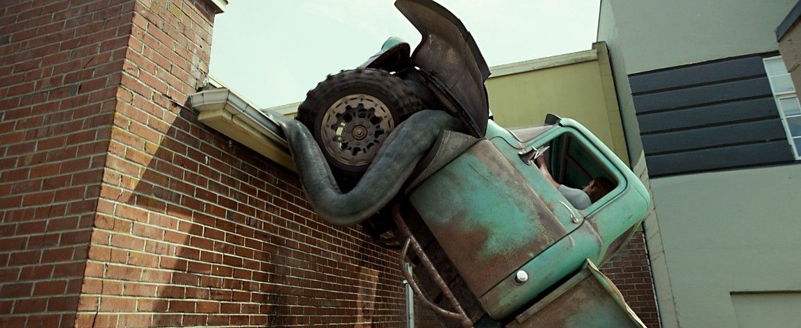 A large snake wrapped around a truck tire, smashing a brick wall. The truck is teal.
