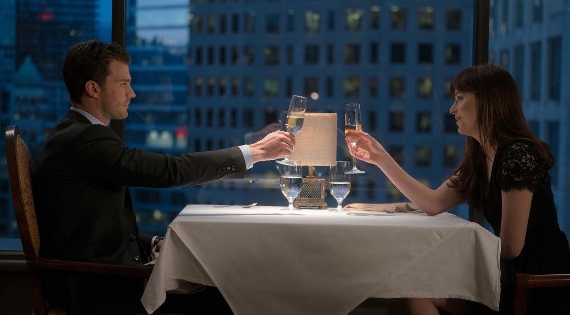 Man and woman toasting champagne at a fancy restaurant, city skyline visible.