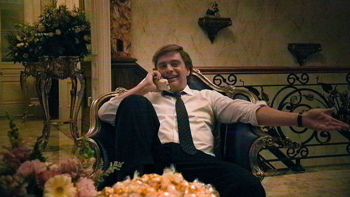 Man in a white shirt and tie, reclining in an ornate chair, talking on a landline, gesturing.