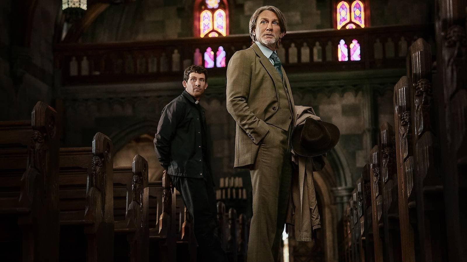 Two men stand in a dark, wood-paneled hall with stained-glass windows and rows of chairs.