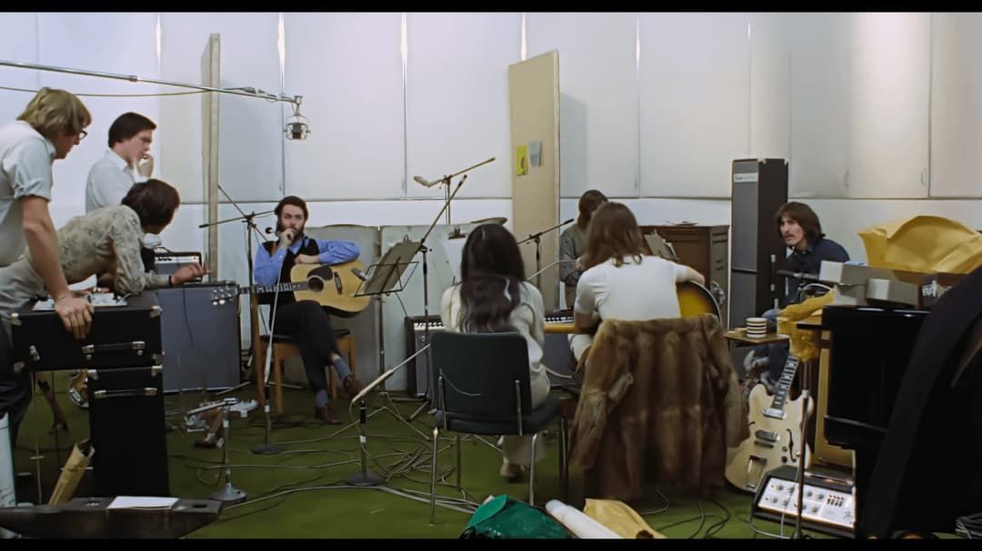 People seated in a music rehearsal studio with instruments, microphones, and equipment scattered around.