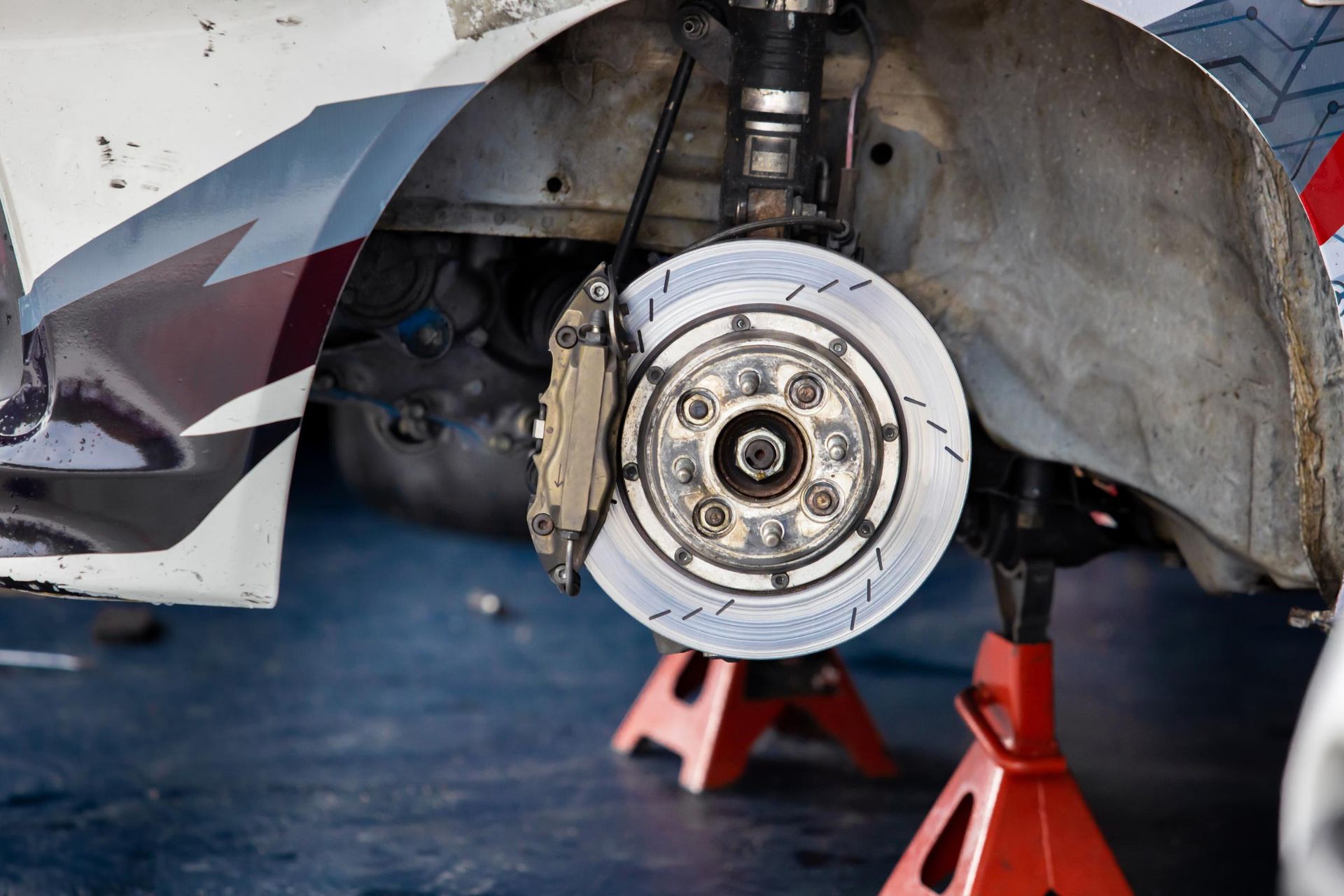 Car with front wheel removed, exposing brake rotor and suspension on jack stands in a garage