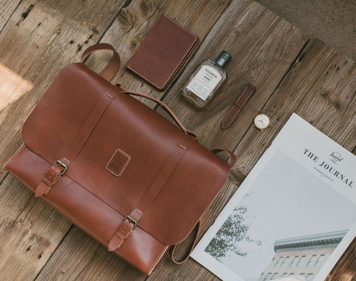 A brown leather satchel backpack, passport, watch, cologne, and magazine arranged on a wooden surface.