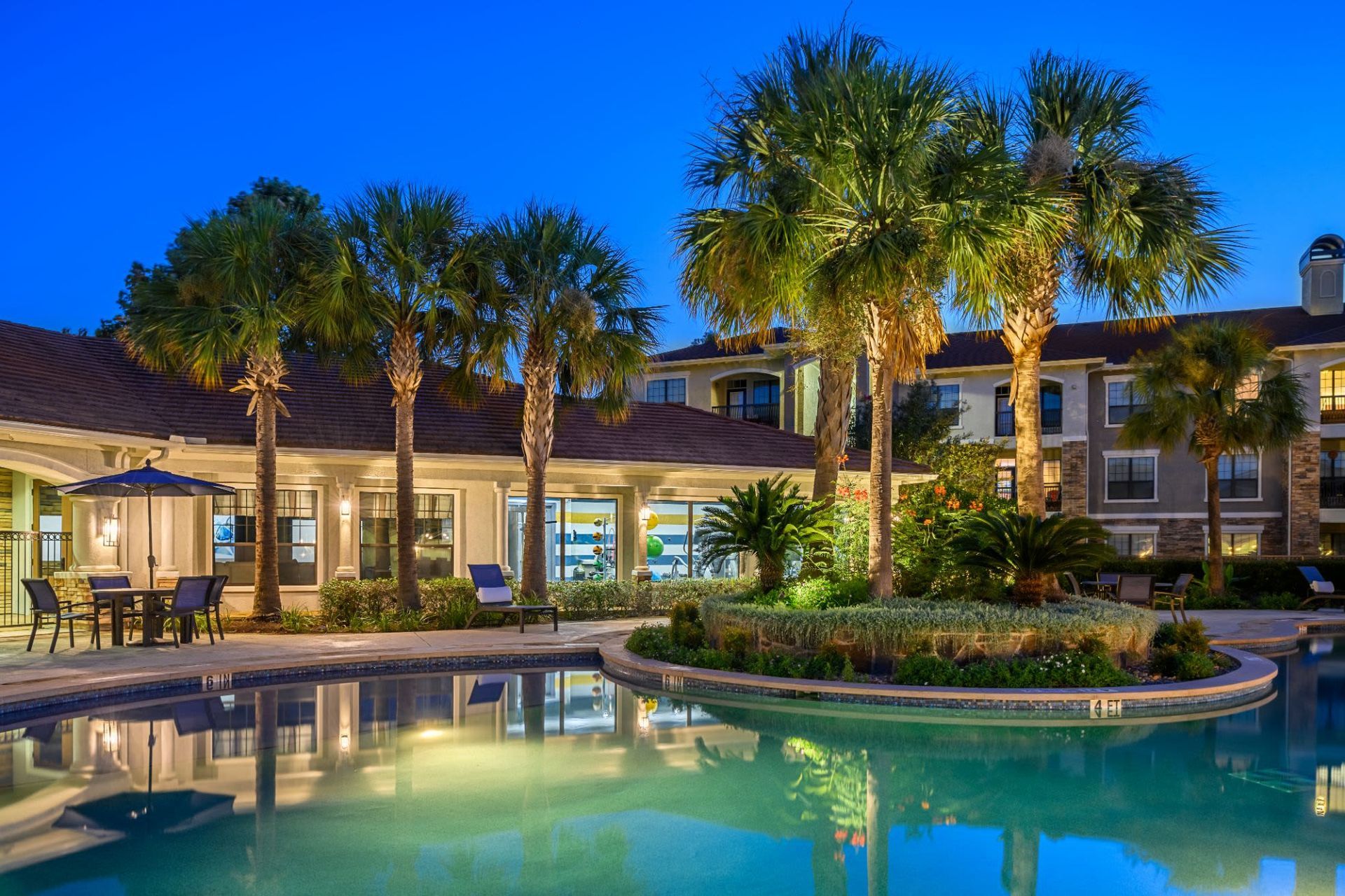 A large swimming pool surrounded by palm trees in front of a building at night at Marquis Lakeline Station in Austin, TX.
