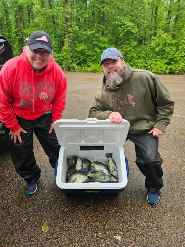 Two people kneel beside a cooler full of fish on a wet road in a wooded area.