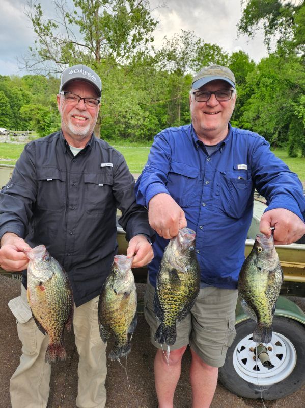 Two anglers holding large fish in front of a trailer, smiling outdoors under cloudy skies
