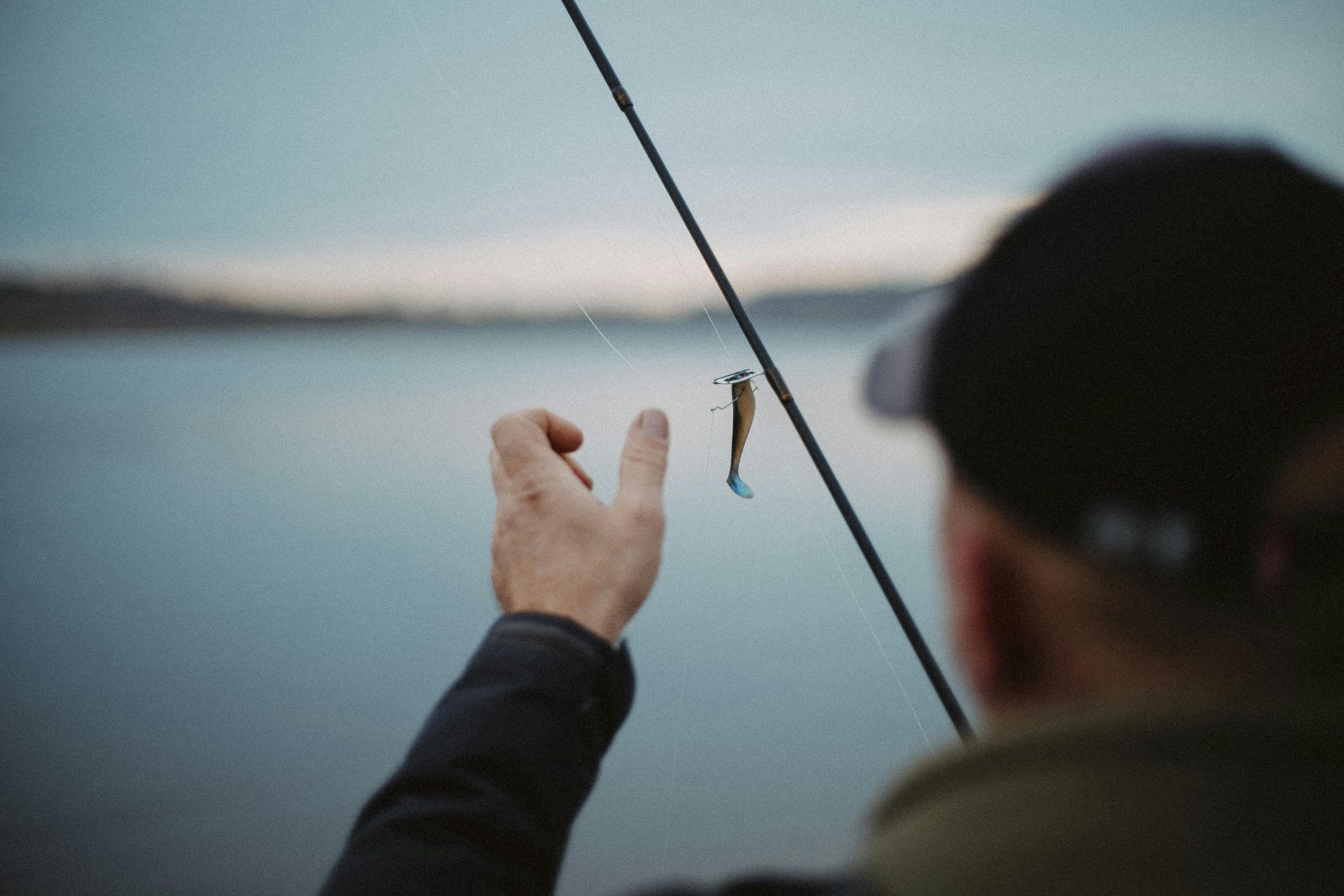 Person fishing by a calm lake, holding a small fish on a line at sunset.