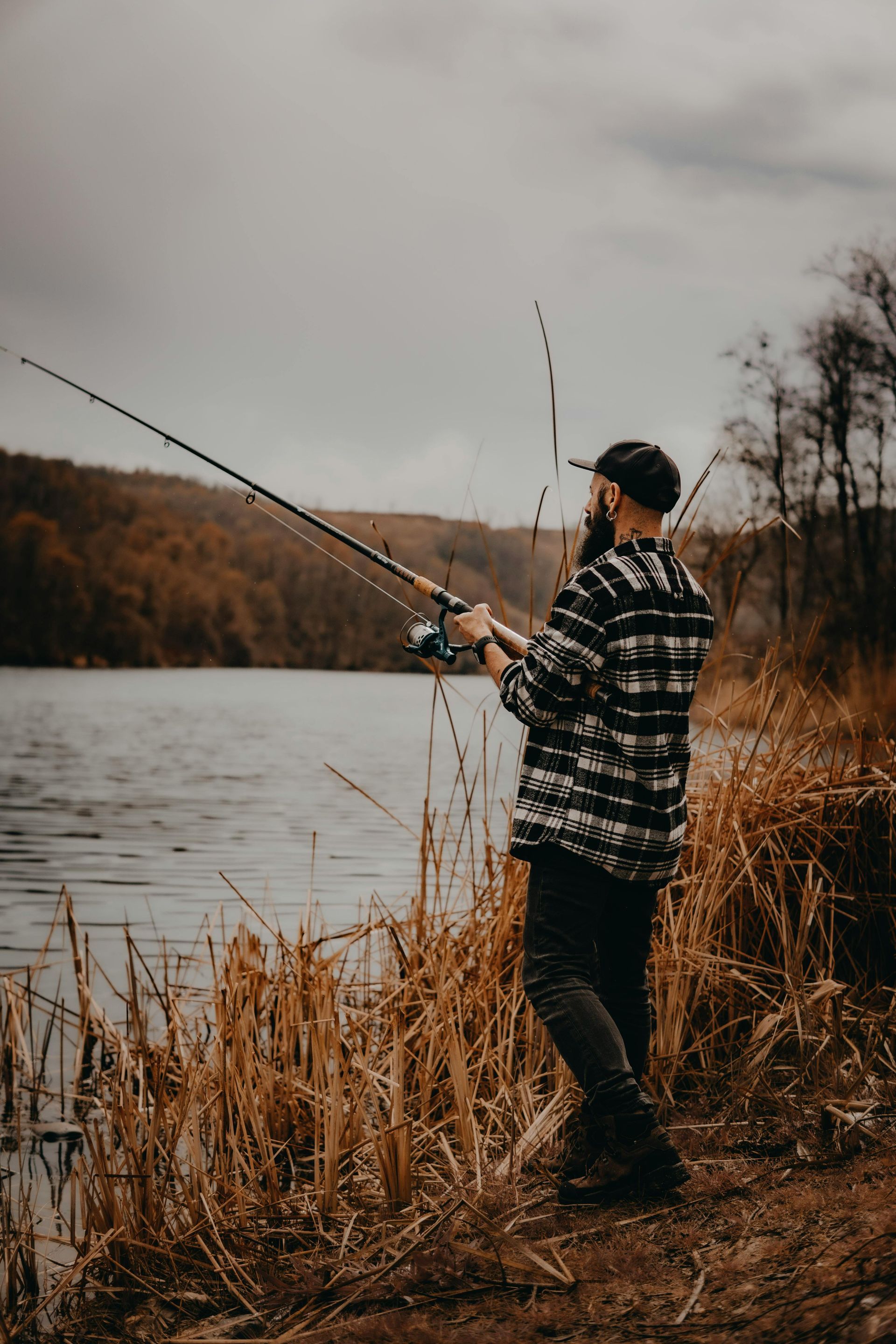 Person fishing by a lakeshore in a plaid jacket on a cloudy day