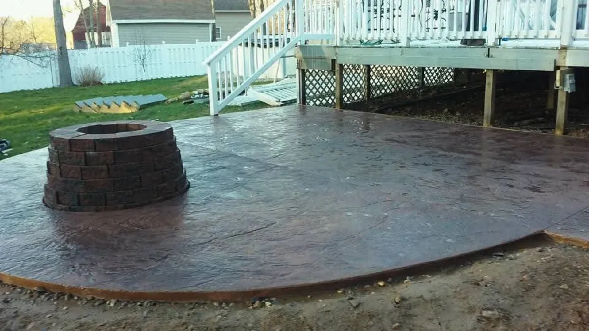 Brick fire pit on a stamped concrete patio beside a house deck and white stairs.