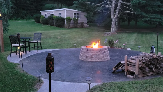 Backyard patio with a lit fire pit, chairs, stacked firewood, and a small shed in the background