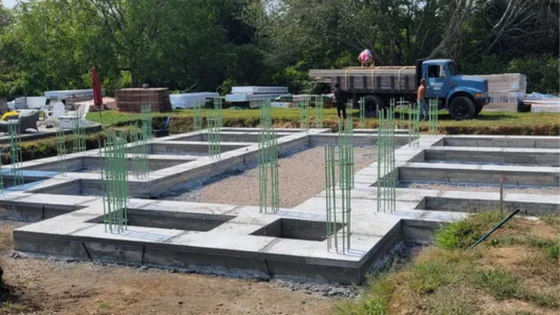 Concrete foundation under construction with rebar columns, and a worker near a truck in the background.