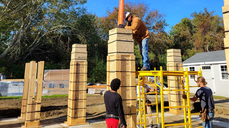 Workers assembling a large wooden structure outdoors beside a yellow lift and a small building.