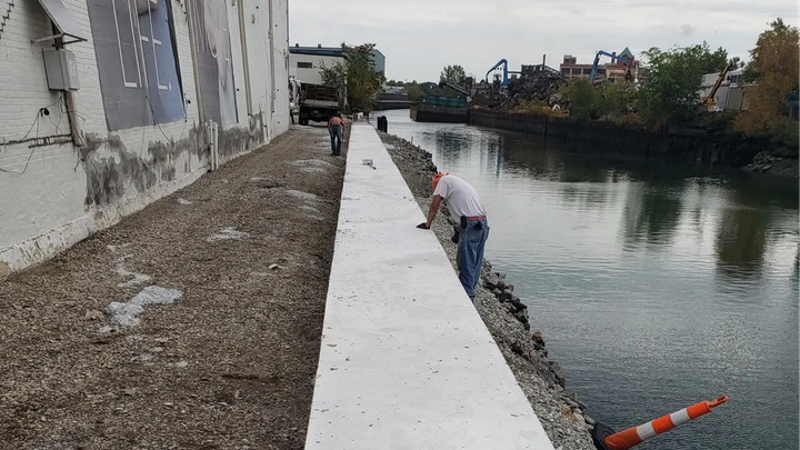 Workers building a white concrete embankment along a canal beside a gravel path and buildings