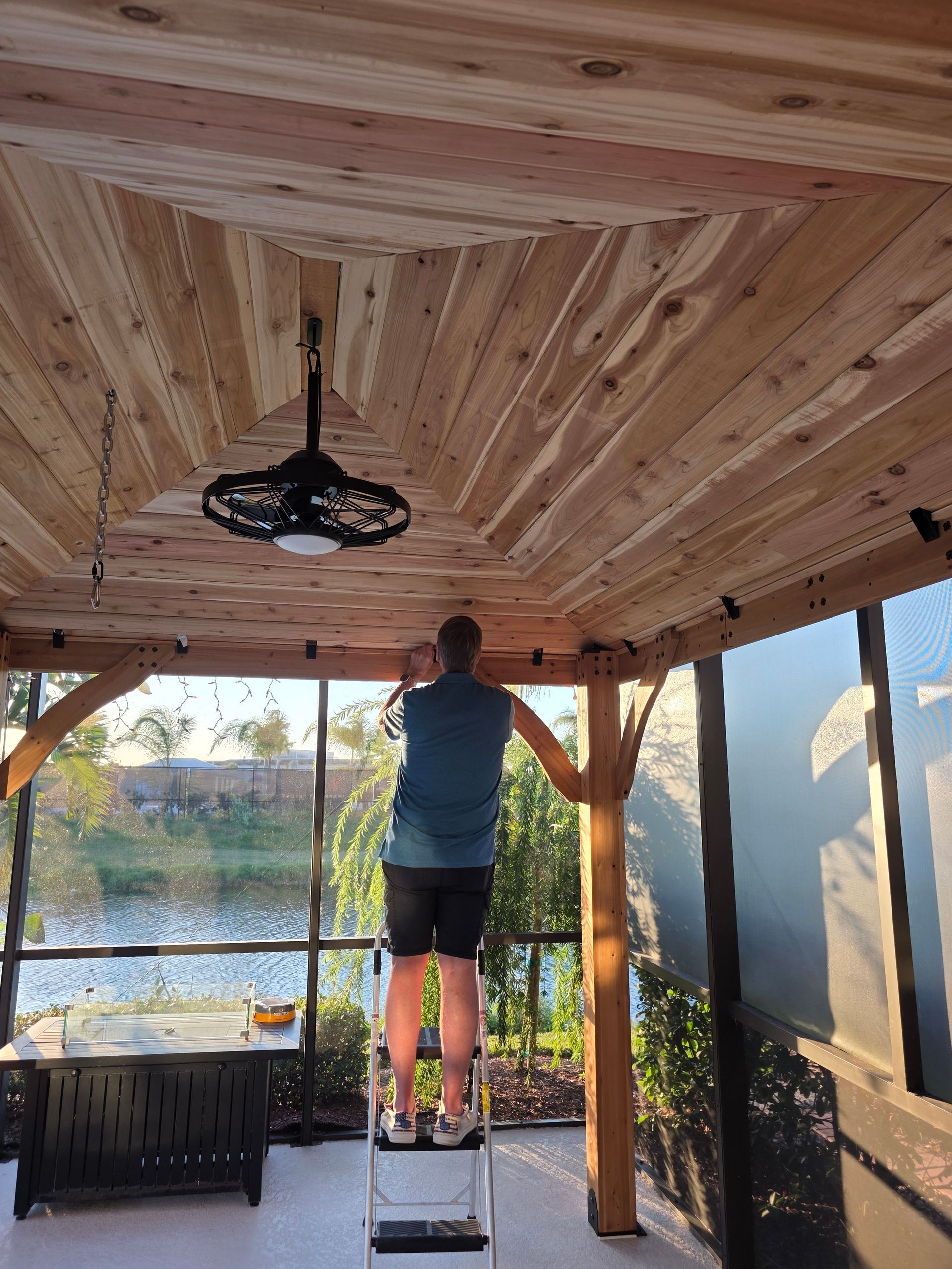 Man on stepladder installing ceiling fan in a gazebo with wood ceiling panels.