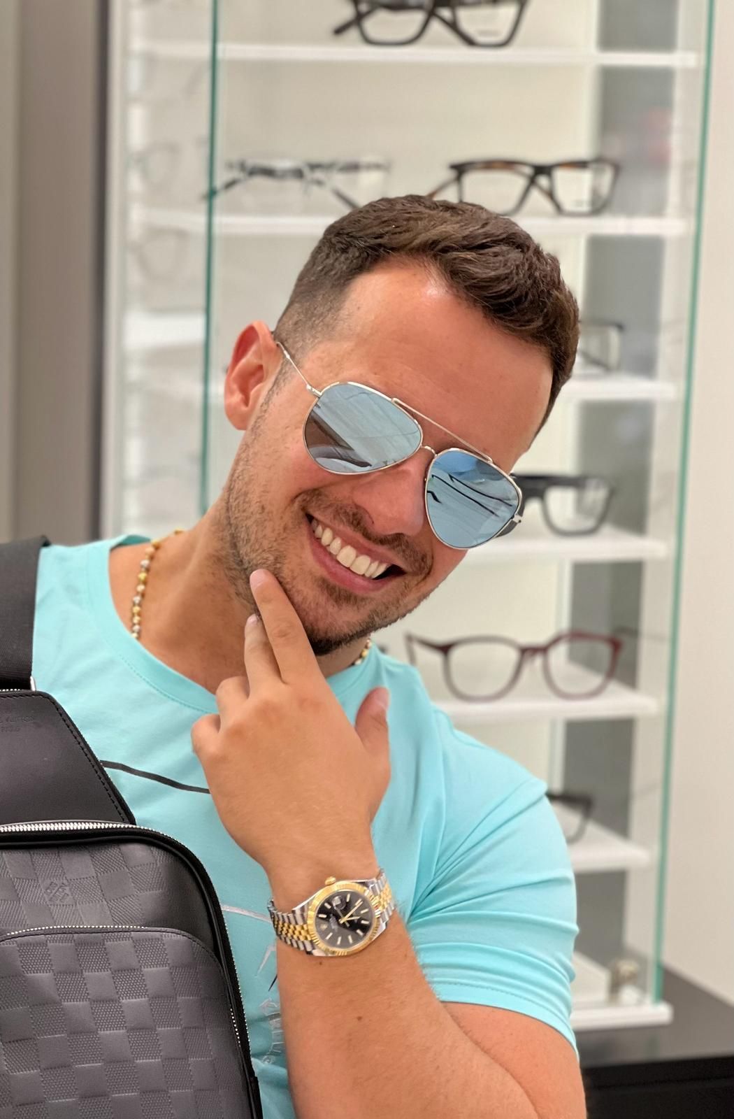 A man wearing sunglasses and a watch is smiling in front of a display of glasses.