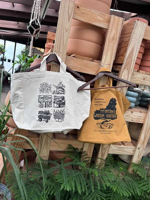 Two tote bags with printed designs hang on a wooden display in a plant-filled market stall.