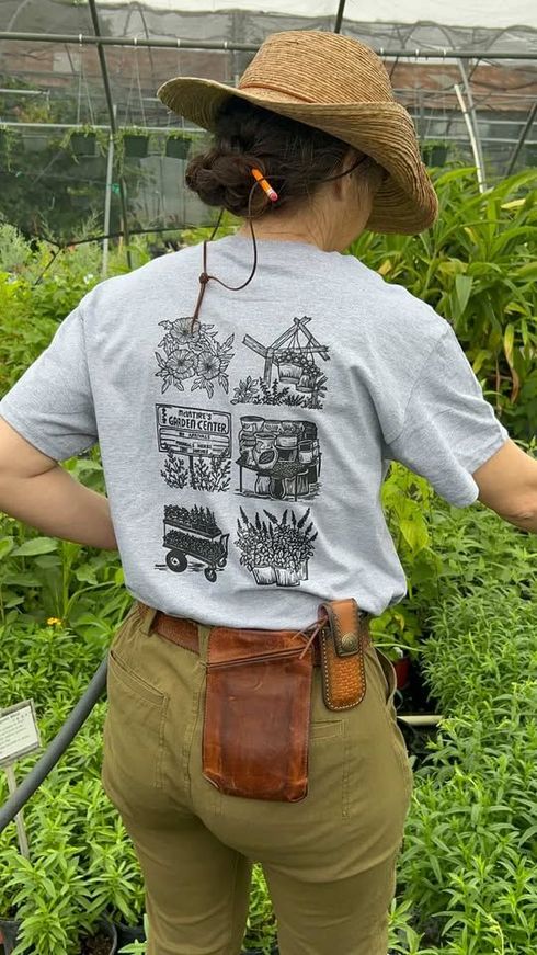 Person in straw hat and gray T-shirt stands in a greenhouse with hands on hips, back facing camera.