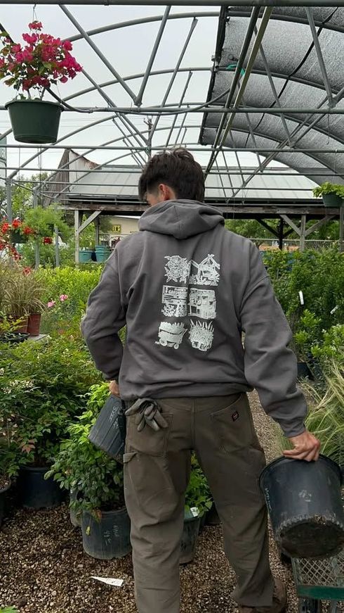 Person in a greenhouse carrying a bucket beside potted plants and flowers