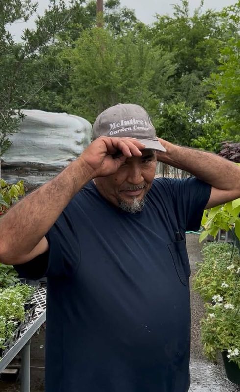 Man adjusting his gray cap in a garden with potted plants and greenery behind him