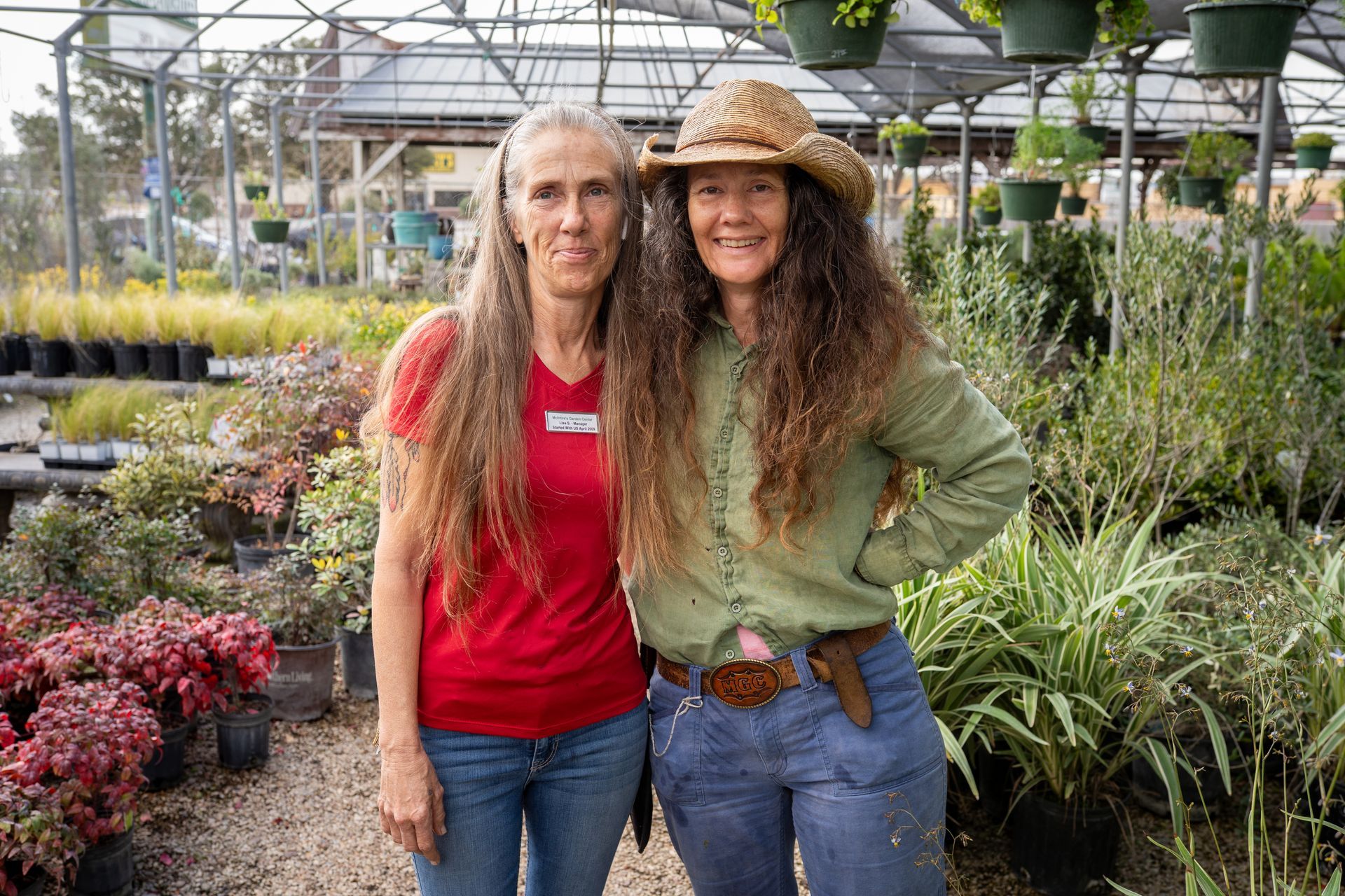 Two women smiling in a greenhouse, standing beside rows of potted plants.